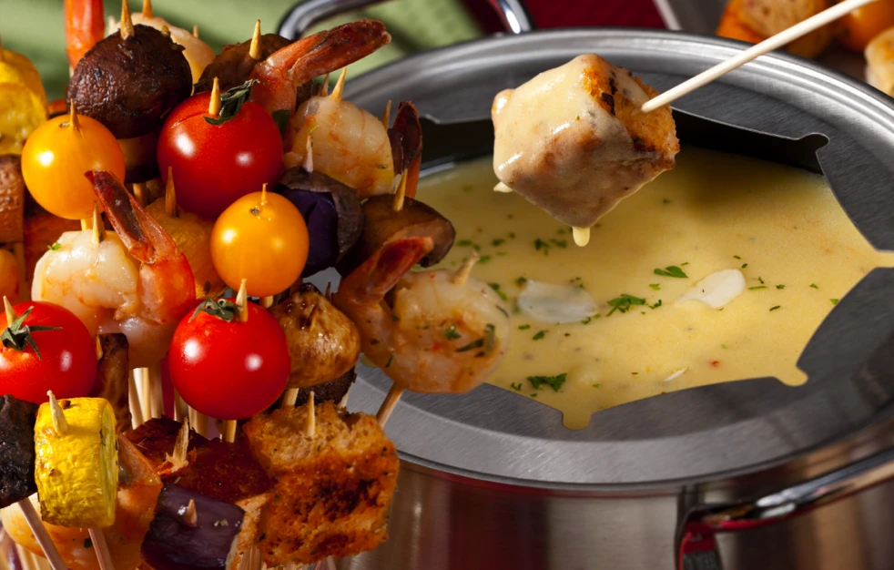 Bread cube dipped into cheese fondue beside skewers of shrimp and vegetables