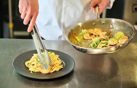 Chef plating buttery pasta with tongs beside a pan of shrimp and greens