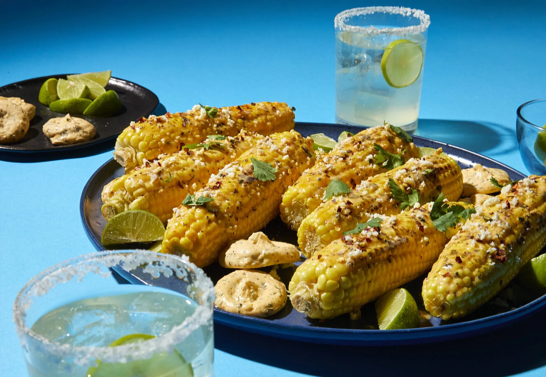 Plate of grilled elote corn served with margaritas on a blue background
