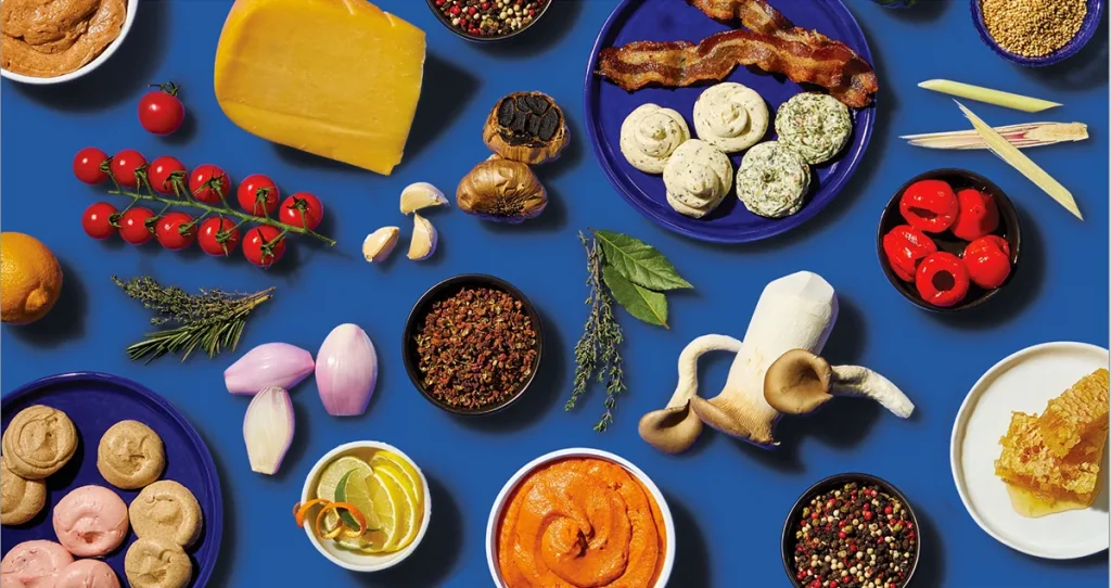 Overhead flat lay of cheese, tomatoes, bacon, spices and herb butter on blue background
