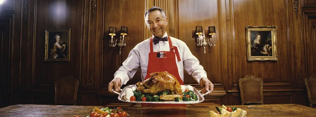 Man in a red apron presenting a roasted turkey in a formal dining room