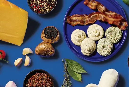 Spices, salts, herbs, sauces and citruses — compound butter medallions on a blue plate surrounded by bacon, aged cheese, garlic, peppercorns and herbs
