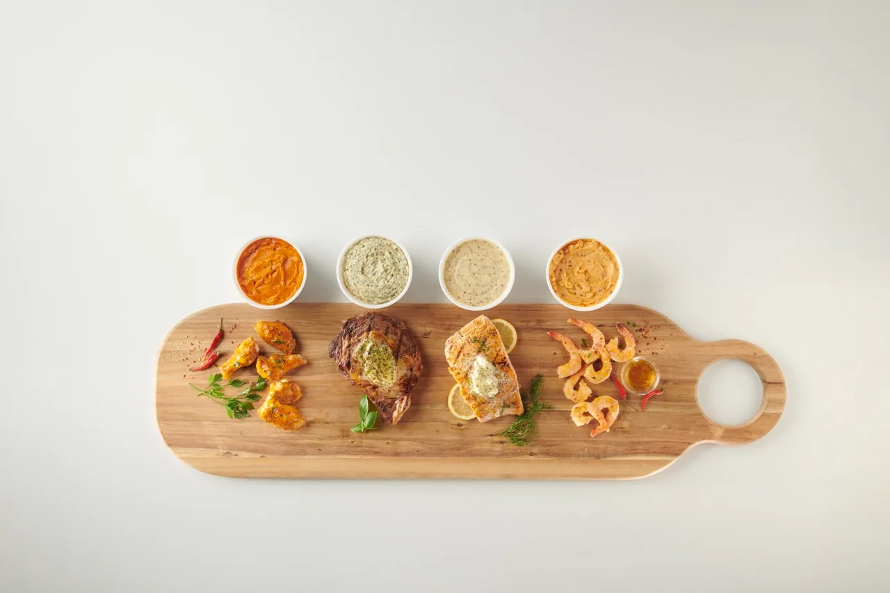 Wood board with seared steak, salmon and shrimp beside four sauce cups