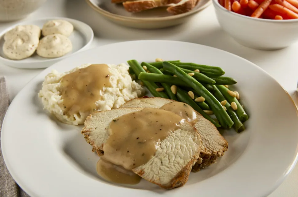 Sliced turkey with gravy, mashed potatoes and green beans beside a plate of seasoned butter dollops