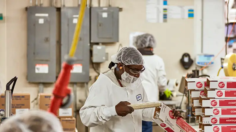 Worker in a hairnet writing on a box amid production floor equipment