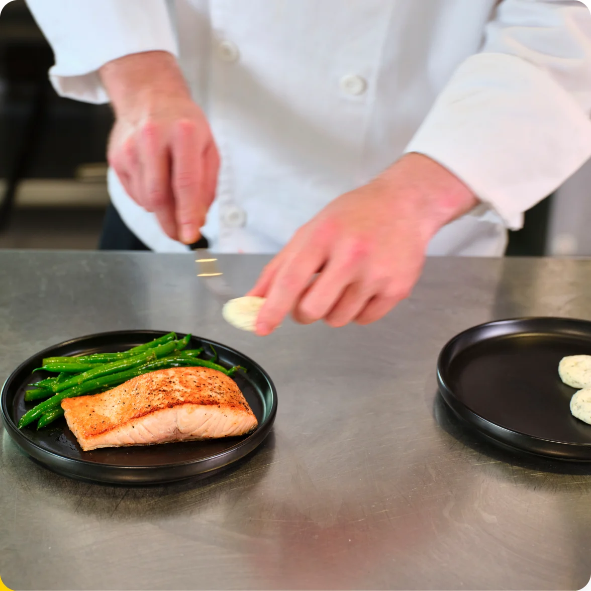 A chef placing a flavored butter dollop onto a plated salmon fillet with green beans