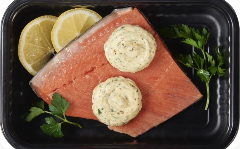 A packaged salmon fillet topped with two flavored butter dollops, lemon and parsley